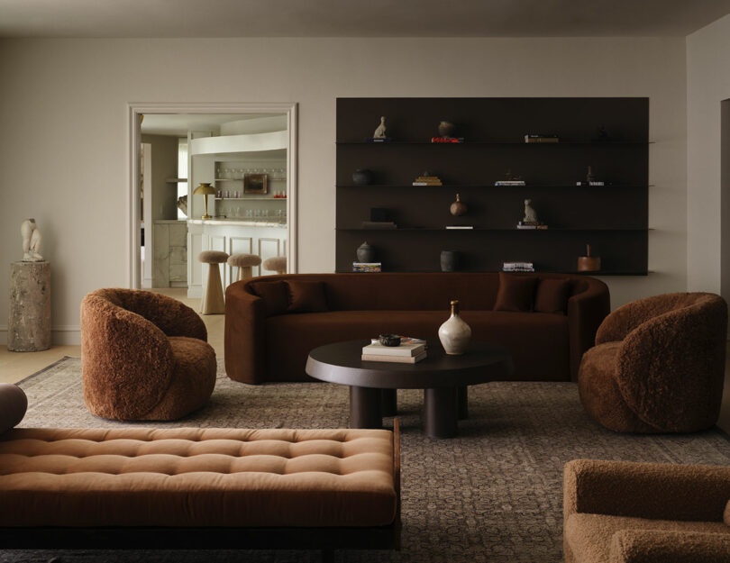 Modern living room with OSKLO-inspired brown curved sofas, a round coffee table, a tan tufted bench, and a wall-mounted shelf displaying small objects; kitchen visible through doorway.