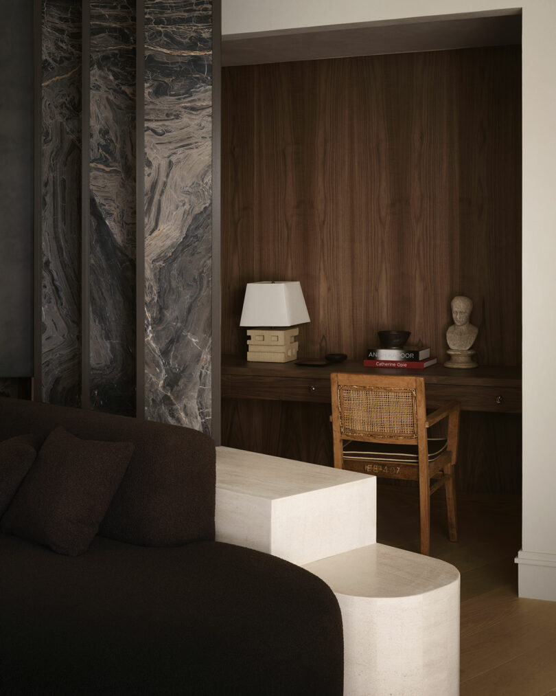A minimalist home office nook with a wooden desk, woven chair, books, decor items, and a boxy table lamp, set against an OSKLO-inspired wood-paneled wall beside a marble divider.