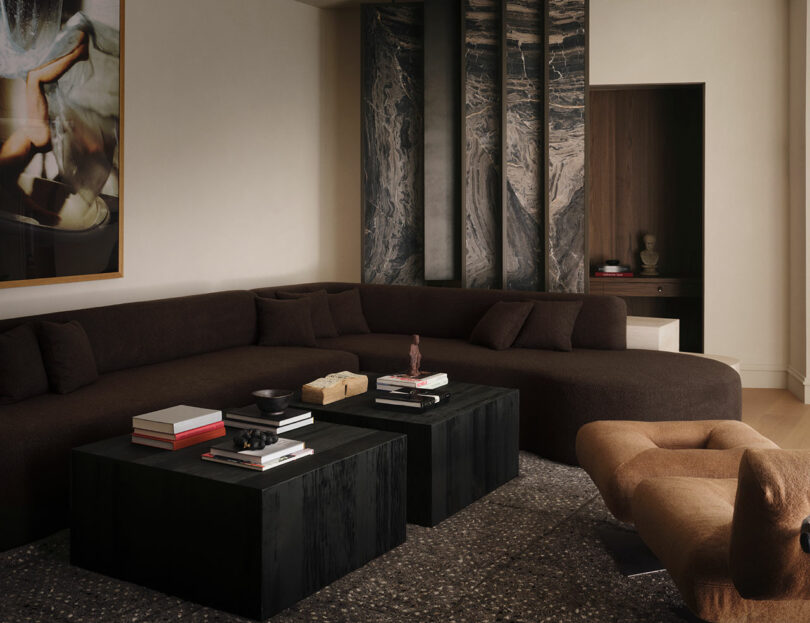 A modern OSKLO living room featuring a dark curved sofa, two black square coffee tables with books and decor, a tan lounge chair, abstract wall art, and striking dark marble panels.