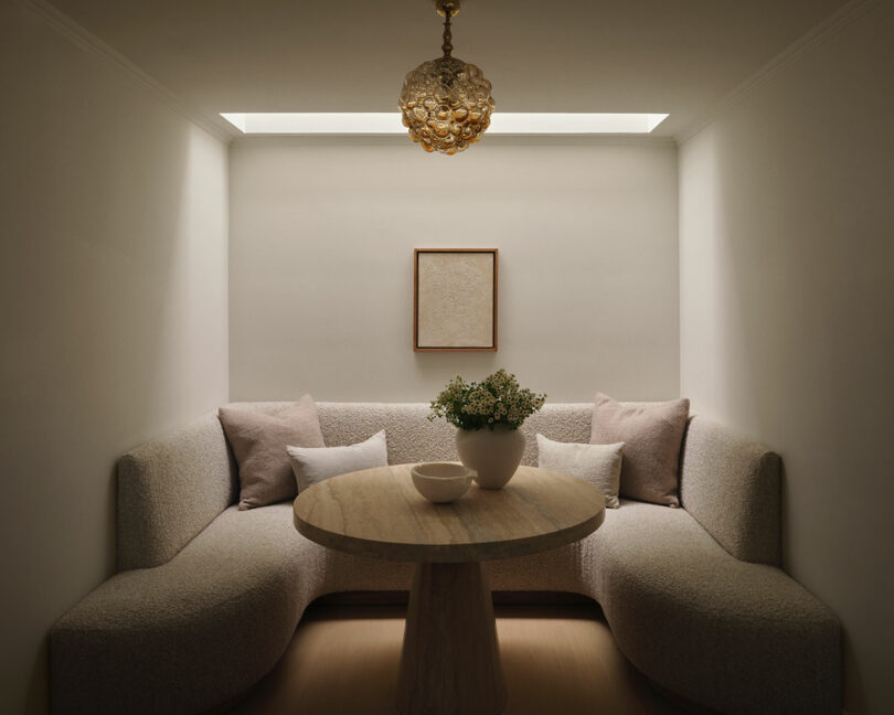 A small, minimalist dining nook by OSKLO features a round stone table, curved sofa seating, a bowl and vase on the table, simple framed artwork, and a decorative ceiling light.