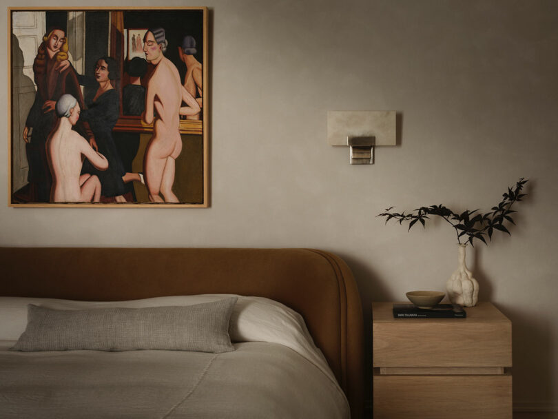 A modern OSKLO bedroom with a brown bed, neutral bedding, a wooden nightstand with books and a vase holding foliage, and a large framed painting of nude figures on the wall.