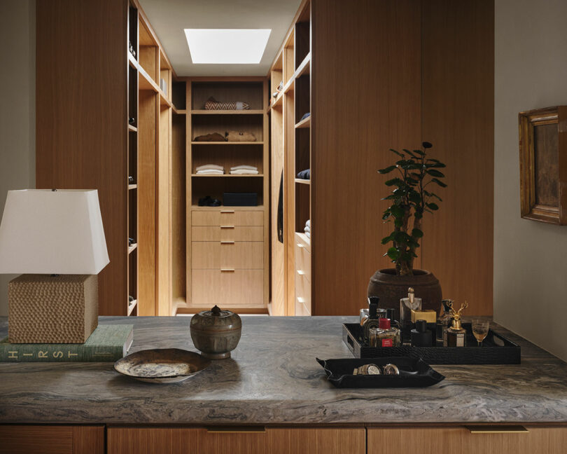 A modern OSKLO walk-in closet with wooden shelves, a marble-topped island, a potted plant, various trays with accessories, a lamp, and stacked clothes visible in the background.