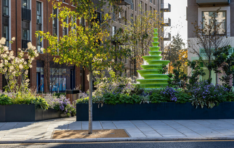 A green tiered fountain stands in a landscaped urban courtyard with trees, plants, and modern apartment buildings in the background.