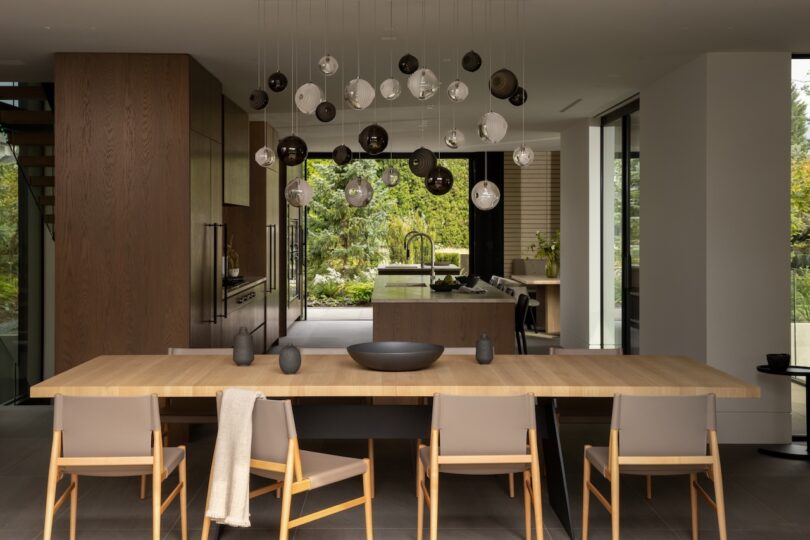 Modern dining area with a long wooden table, neutral chairs, hanging spherical light fixtures, and a view into a contemporary kitchen with large windows showing greenery outside.