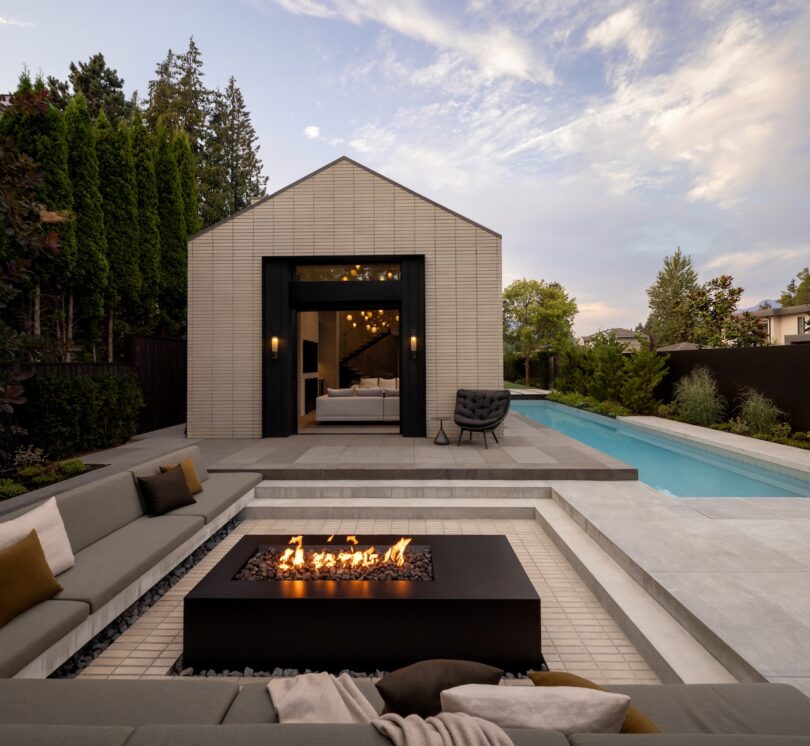 Modern backyard with a rectangular fire pit, gray seating, swimming pool, and a contemporary pool house with large sliding doors.