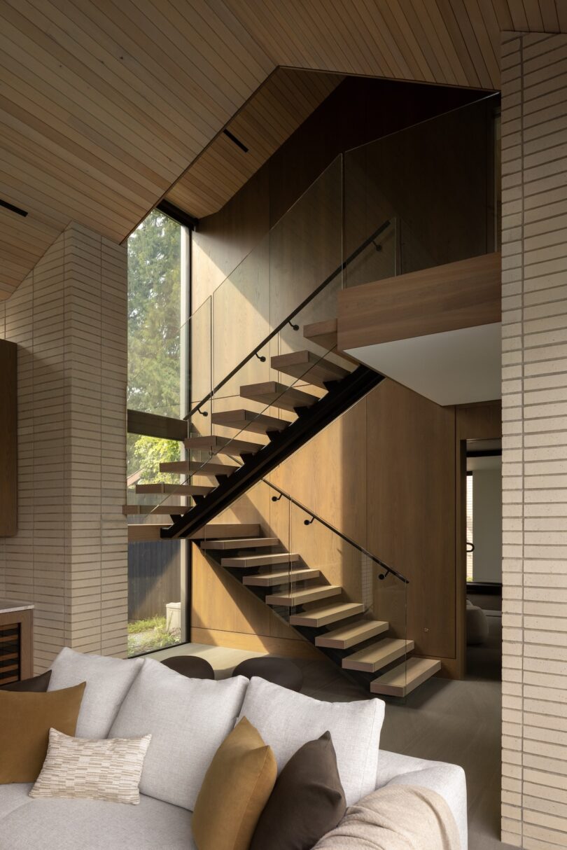 Modern staircase with glass railing and wooden steps in a contemporary living room with a beige sofa and large windows.