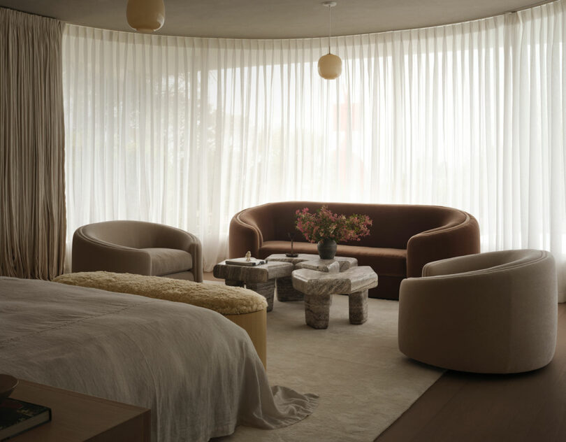A modern OSKLO living area with curved beige chairs, a brown sofa, stone coffee tables, and sheer curtains allowing natural light into the neutral-toned space.