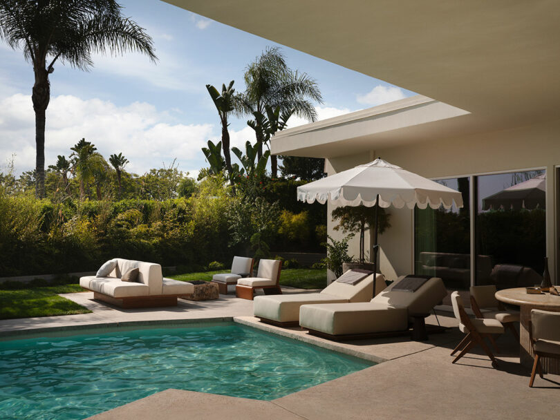A modern backyard patio by OSKLO with a pool, lounge chairs, couches, umbrella, and dining table, surrounded by lush greenery and palm trees.