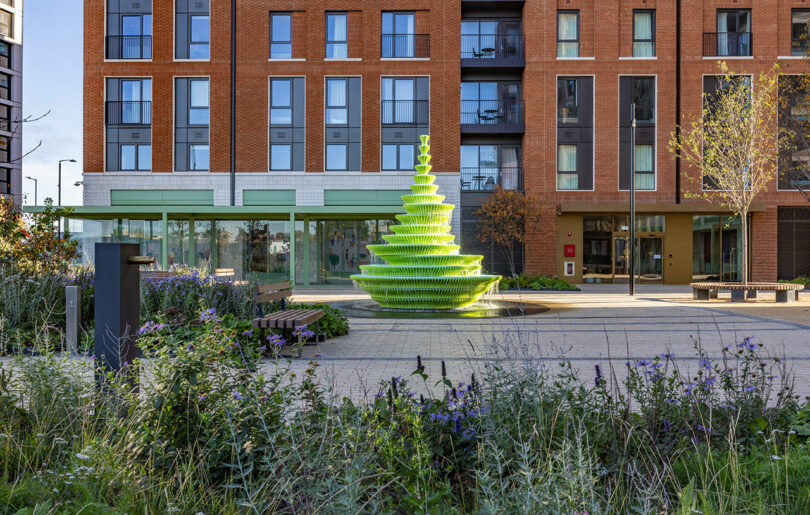 A modern plaza features a tiered, neon green fountain surrounded by benches, plants, and a red brick apartment building in the background.