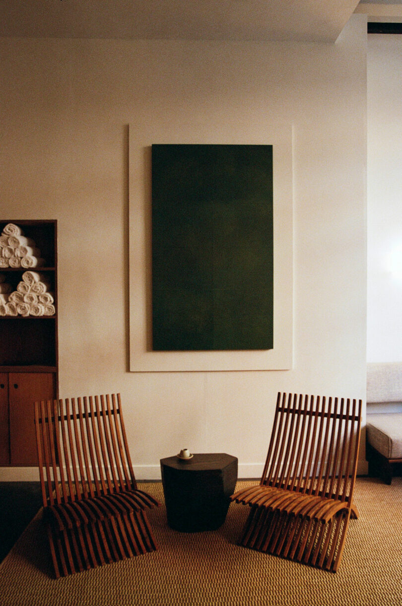Two wooden slat chairs face a small hexagonal table with a cup on it, beneath a large green abstract painting in a minimalist room.
