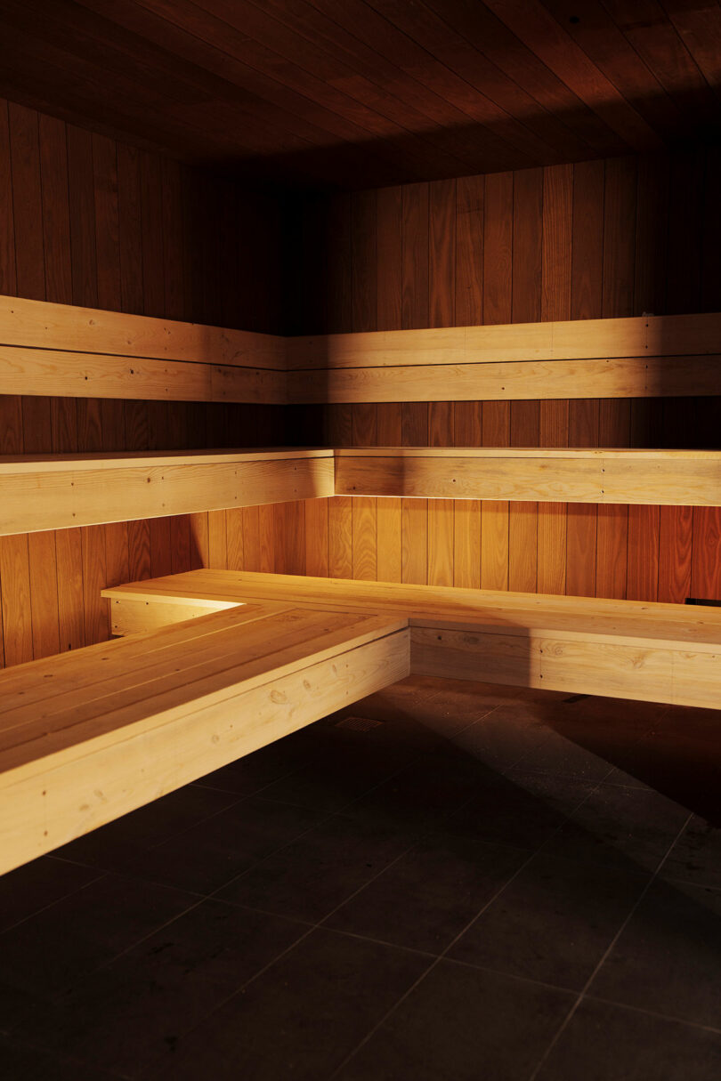 Empty wooden sauna interior with tiered bench seating and dark floor tiles, lit by natural light.