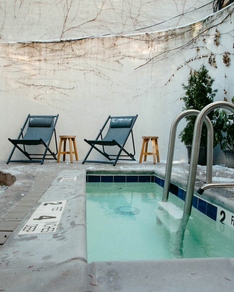 Two empty lounge chairs and stools sit beside a small pool with clear water, set against a white wall with sparse vines and some greenery.