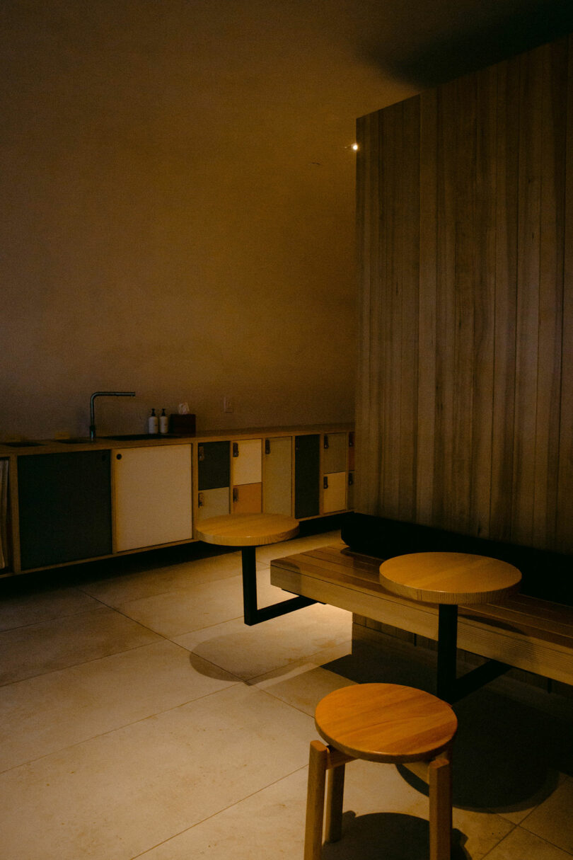 A dimly lit room with a wooden bench, round wooden stools, and a kitchenette with a sink and bottles on the counter.