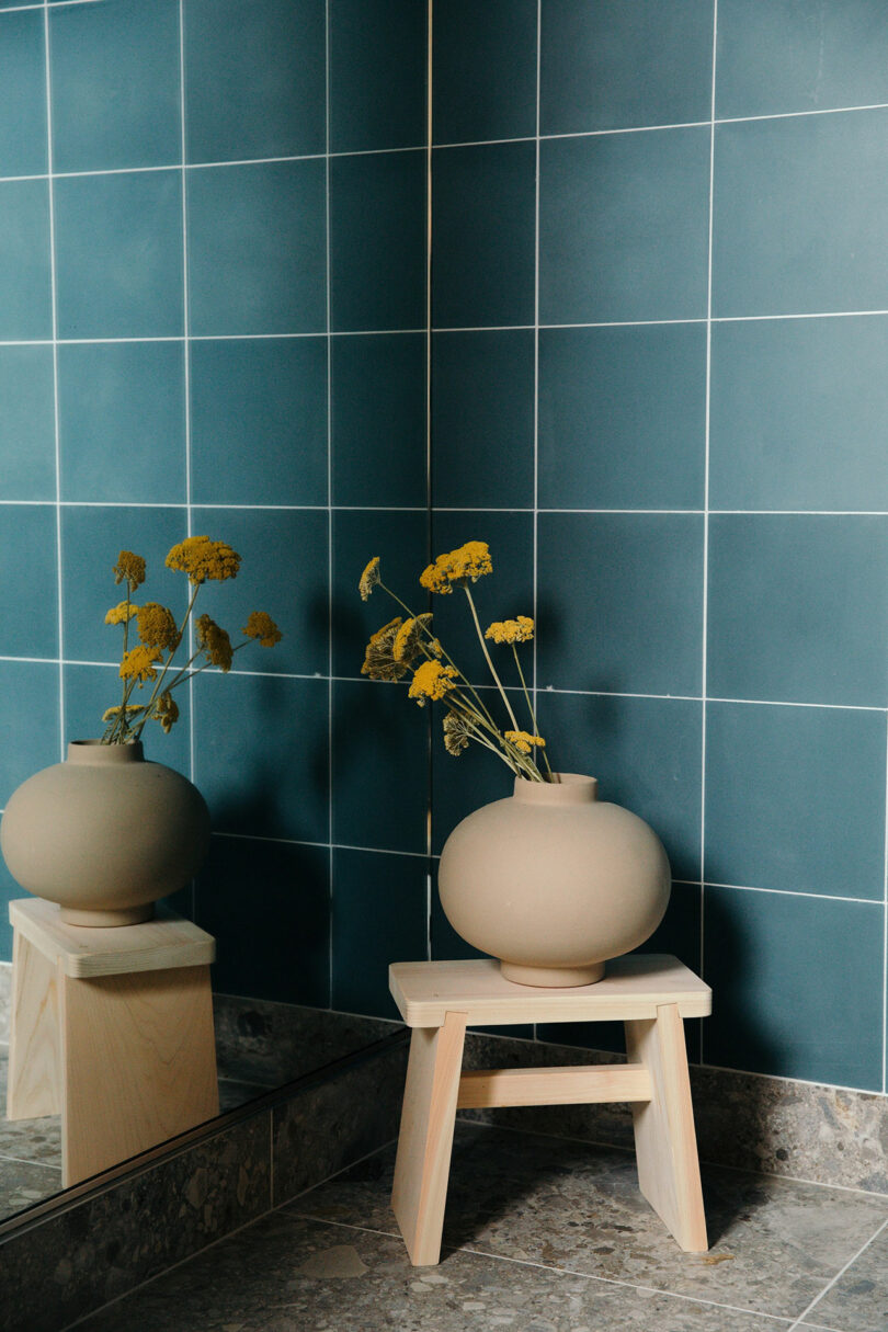 A round beige vase with yellow flowers sits on a small wooden stool in front of a mirrored blue-tiled wall.