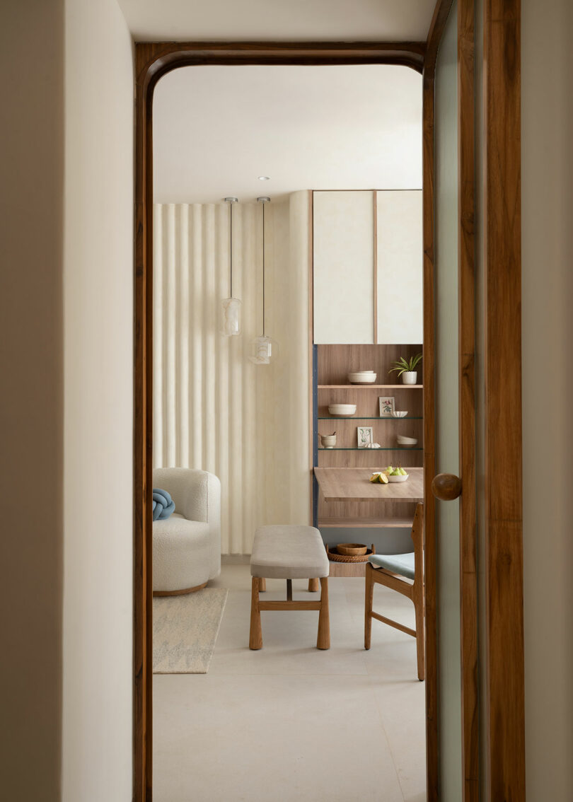 View through a doorway into a minimalist room with a curved white armchair, wooden bench, chair, shelving with decor items, and neutral-toned walls.