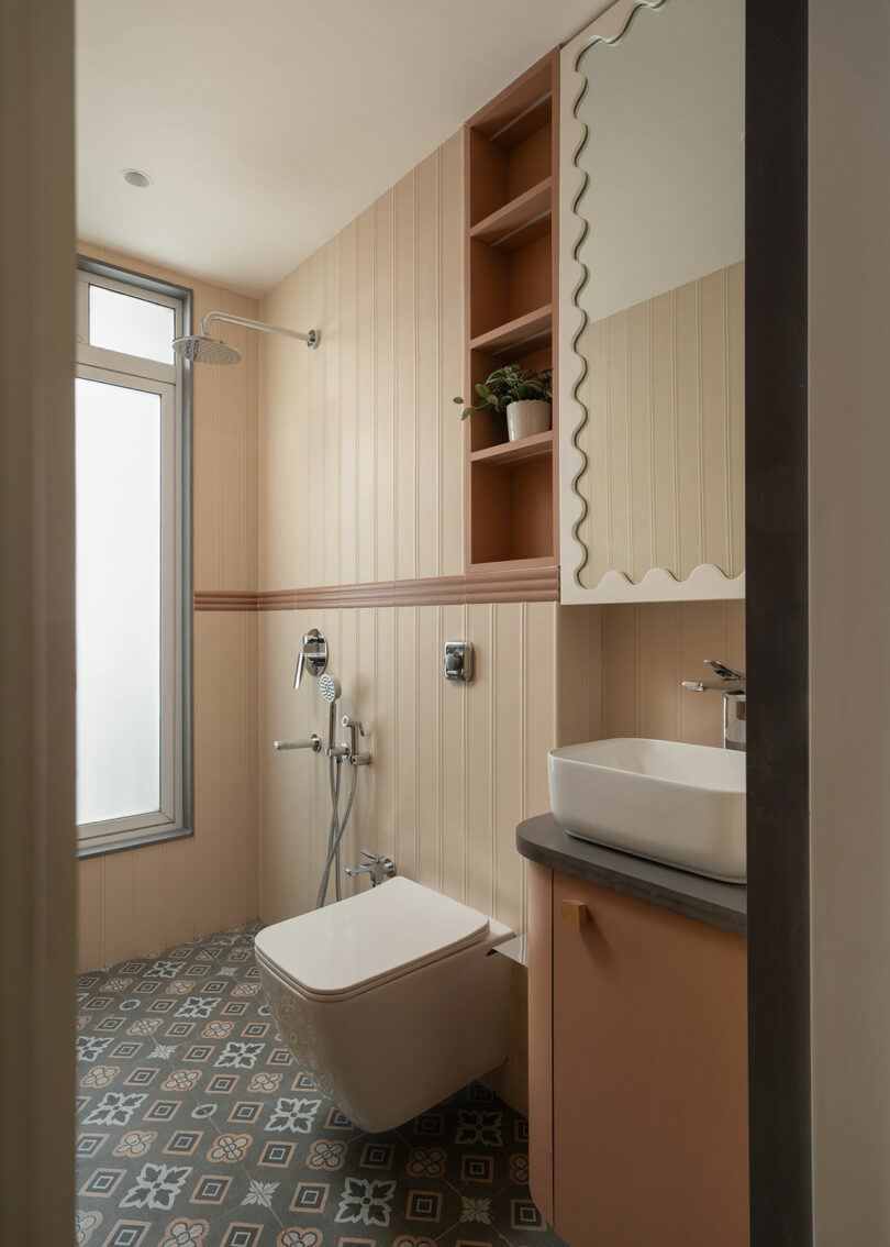 Modern bathroom with patterned tile floor, wall-mounted toilet, shower, and sink with a rectangular mirror. Built-in shelves hold a plant and other items. Frosted window provides natural light.