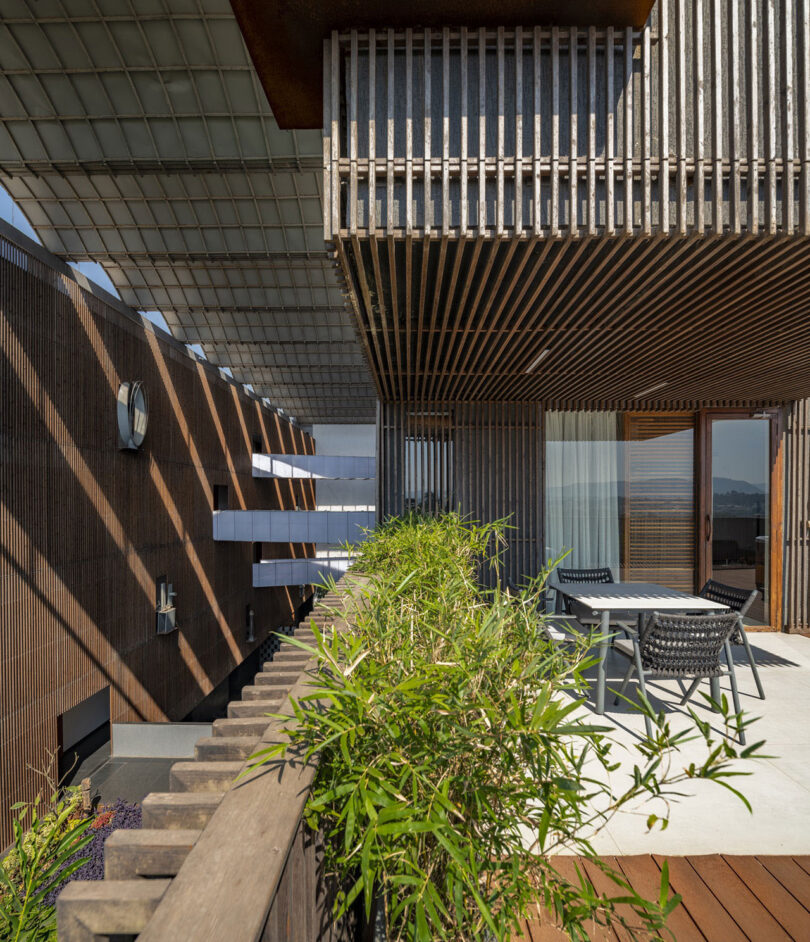 Outdoor terrace by Malik Architecture features wooden slatted walls and ceiling, lush green plants in planters, and a table with four chairs, with large glass doors leading inside.