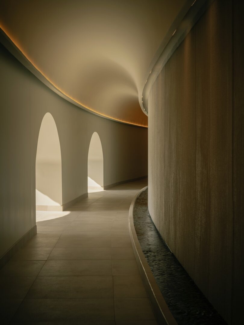 Curved hallway with arched openings on the left and a textured wall with a narrow water feature on the right, softly lit by recessed lighting.