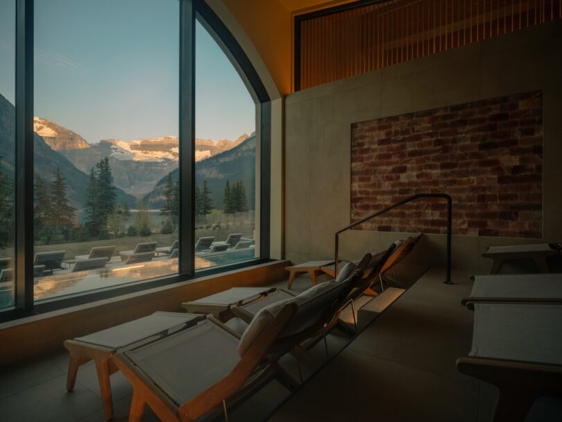 Several lounge chairs face a large window with a mountain view at sunset, inside a modern room with warm lighting and a brick feature wall.