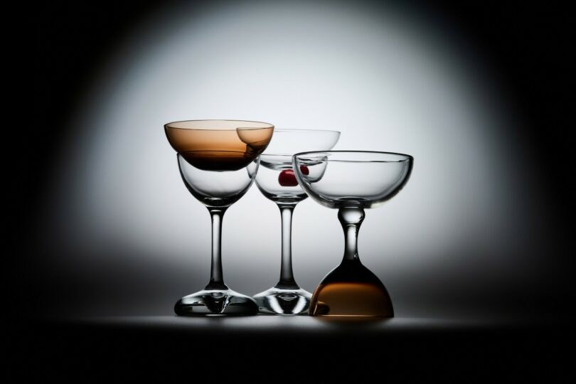 Three coupe glasses are arranged side by side; two are upright, one is upside down. One glass contains a red cherry, and two have a subtle amber tint