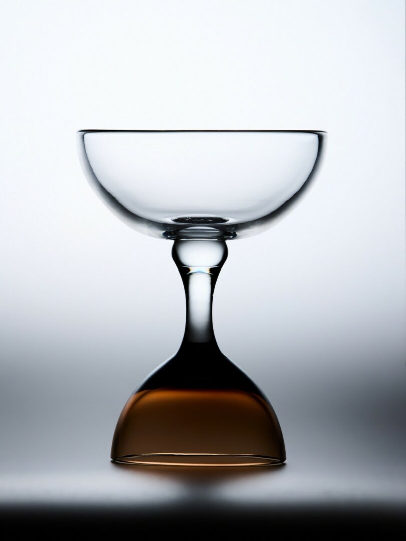A glass coupe is positioned upright atop an inverted amber glass, creating a symmetrical, double-stemmed appearance against a white background