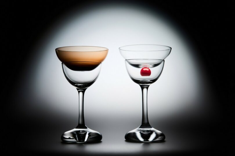 Two empty cocktail glasses on a white and black gradient background; one glass has a brown tint, the other contains a single red cherry
