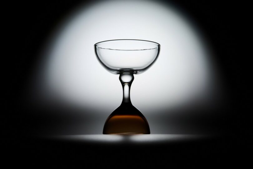 A clear cocktail glass is photographed upside down against a white and black gradient background, with a shadow and light highlighting its shape