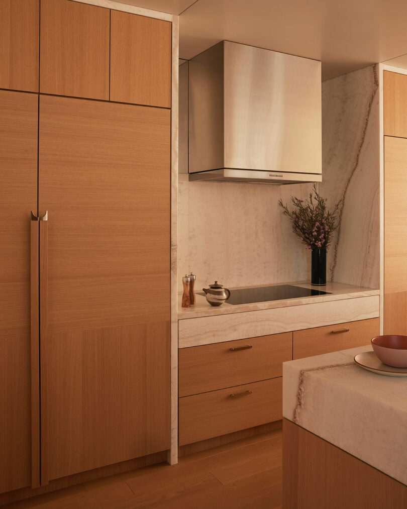 A modern kitchen with light wood cabinets, built-in refrigerator, marble countertops and backsplash, induction cooktop, stainless steel hood, and a vase with flowers.