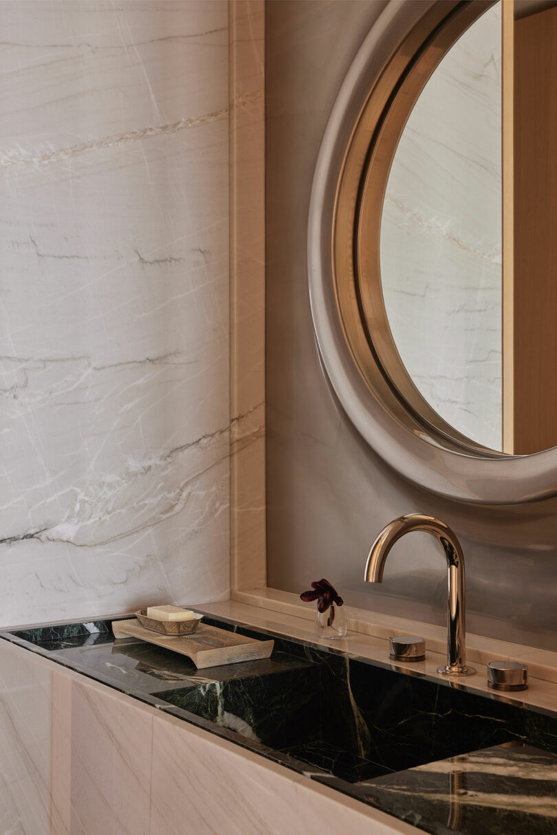 Modern bathroom sink with a marble countertop, oval wall mirror with gold frame, and a gold faucet, set against marble-patterned walls.