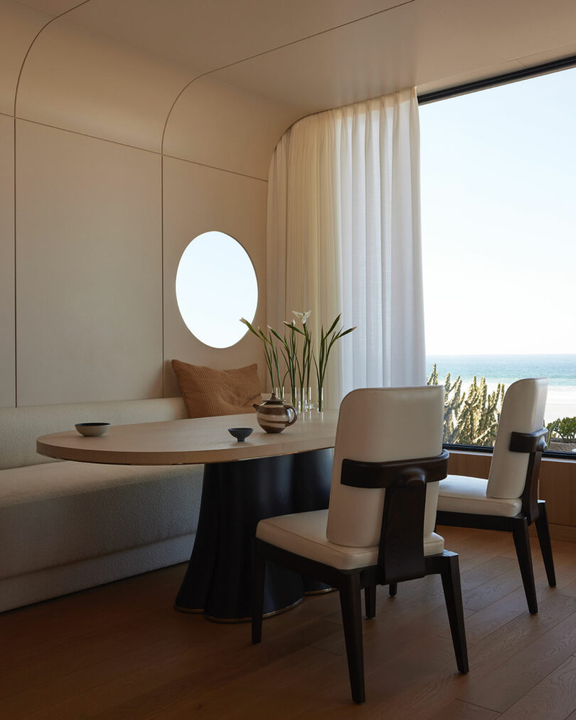 A modern dining area with a round table, white chairs, a built-in bench, a round window, and a large window with white curtains overlooking the ocean.