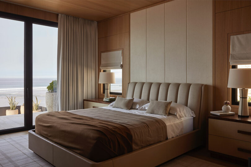 A modern bedroom with a large upholstered bed, beige bedding, bedside tables with lamps, and floor-to-ceiling windows showing a view of the ocean.