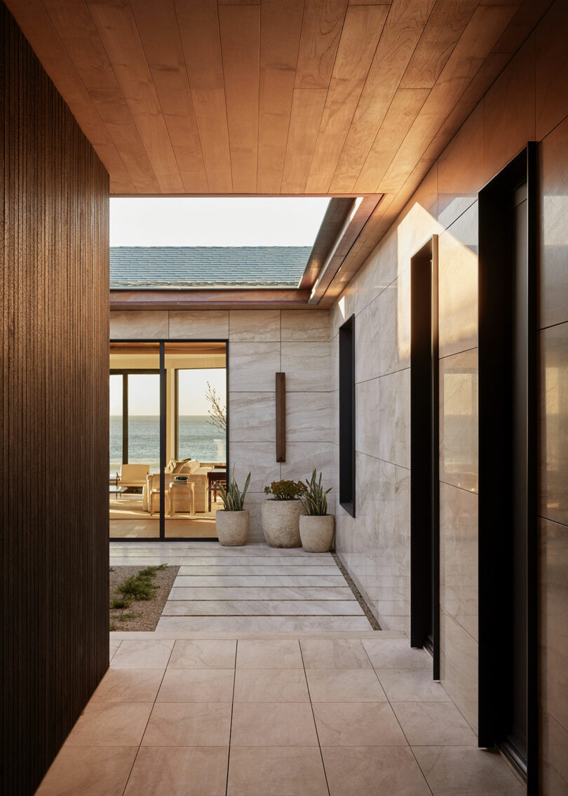 Hallway with light wood ceiling and tiled floor leads to a patio with potted plants and a view of the ocean through glass doors. Modern, minimalist architecture.
