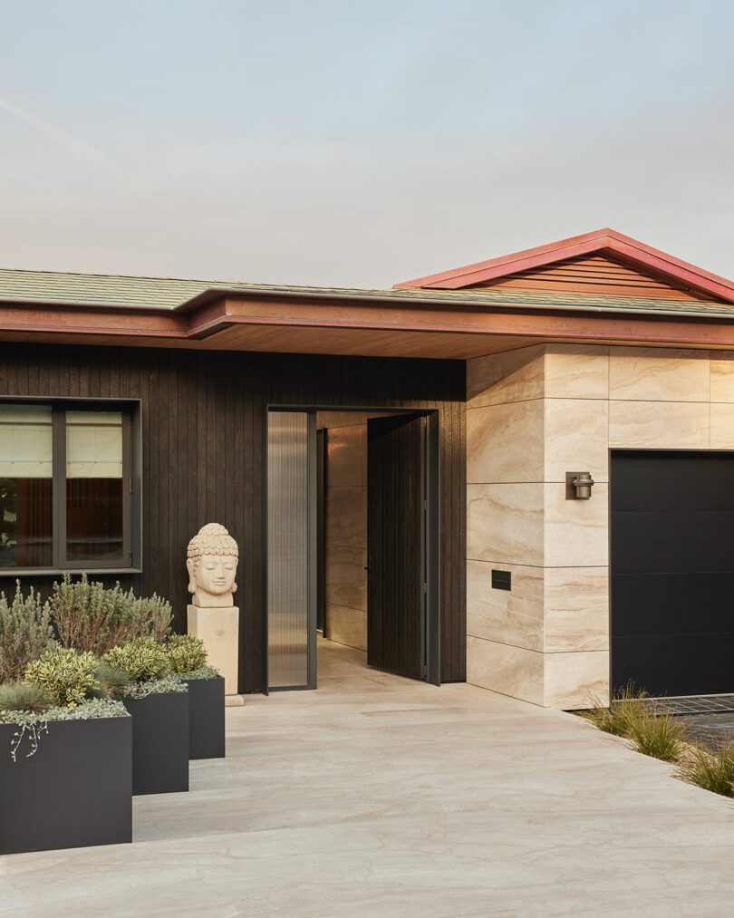 Modern house exterior with black and beige walls, large potted plants, a Buddha head statue by the entrance, and a partially open glass door.