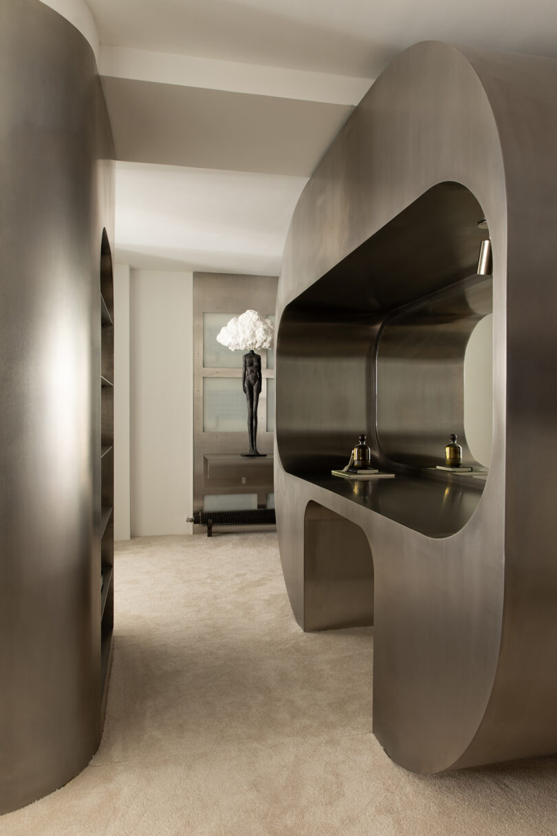 A modern interior with curved metallic structures, beige carpet, a desk with two small sculptures, and a statue with white flowers in the background.