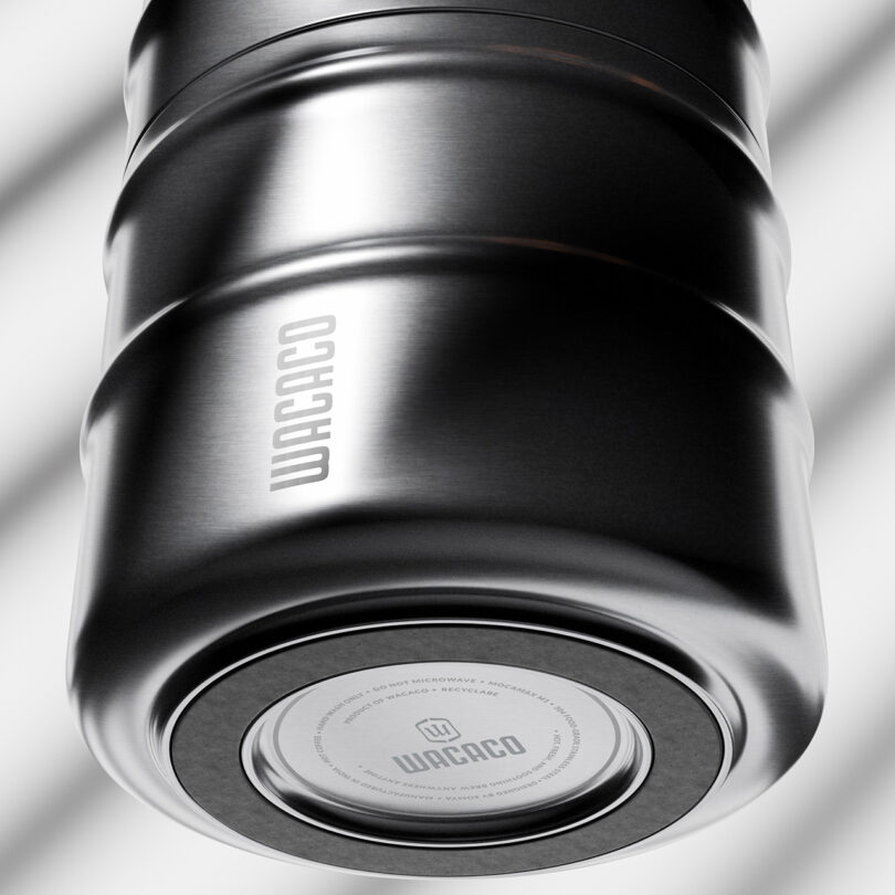 Close-up view of a metallic Wacaco travel mug, showing the brand name on the side and bottom, with a blurred background featuring diagonal lines.