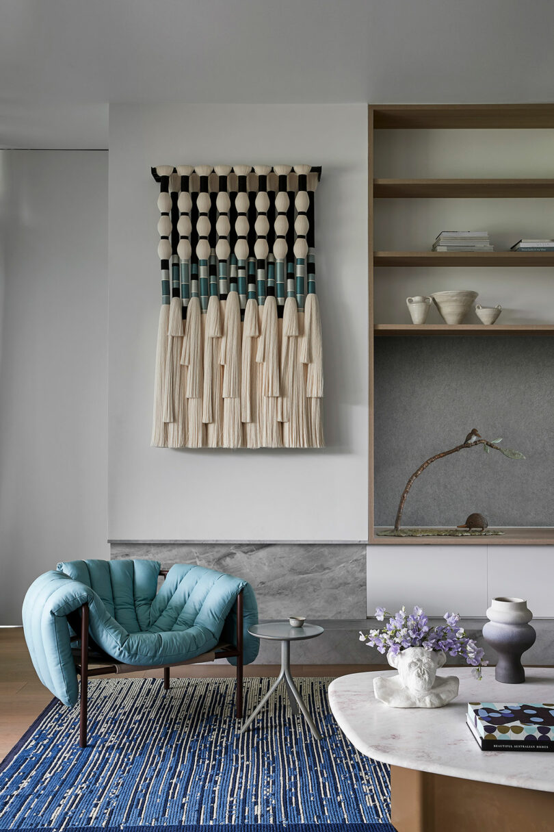 Modern living room with a blue lounge chair, round side table, blue rug, wall hanging textile art, open shelving, and a coffee table with flowers and books.