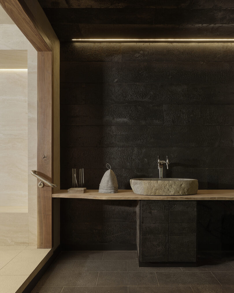 Minimalist bathroom with dark textured walls, a wooden countertop, stone sink, and modern fixtures, illuminated by recessed lighting.