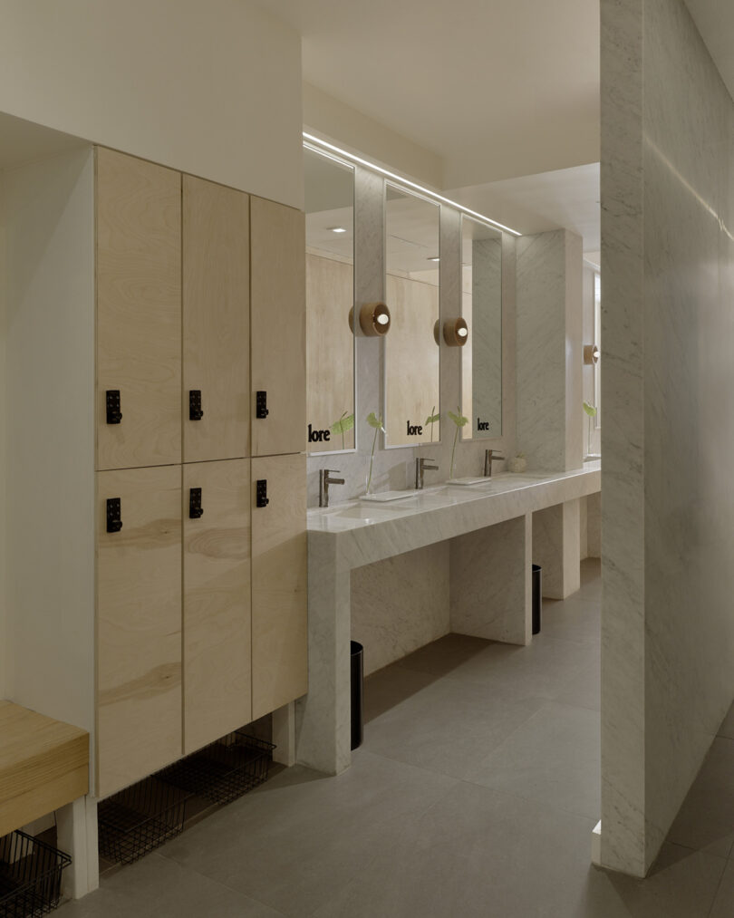 Modern locker room area featuring light wood lockers, a marble countertop with three sinks and mirrors, and neutral-colored walls and flooring.