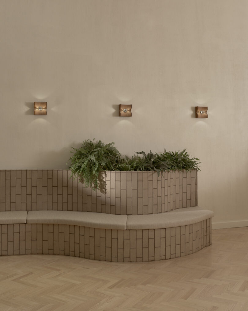 Curved brick bench with beige cushion features built-in planter filled with ferns; three small wall lights above against a plain beige wall.