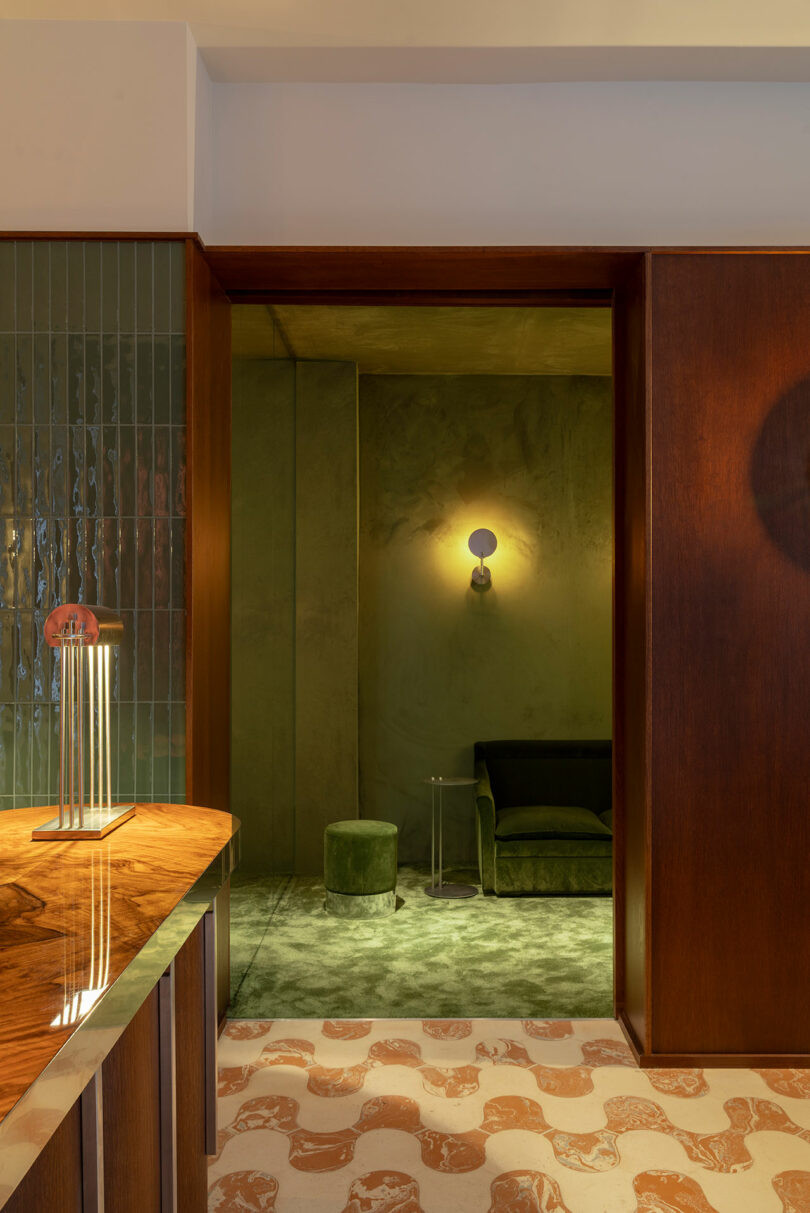 A modern interior with a glossy wood counter, patterned floor, green tiled wall, and a green room with a sofa, ottoman, and wall sconce in the background.