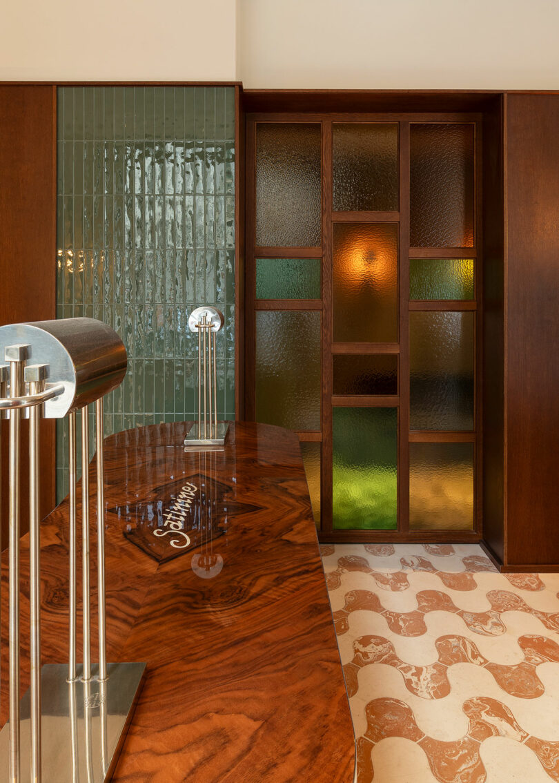 Reception desk with a glossy wood surface, two metal lamps, green tile wall, and a partition with multicolored frosted glass panels, with patterned floor tiles.