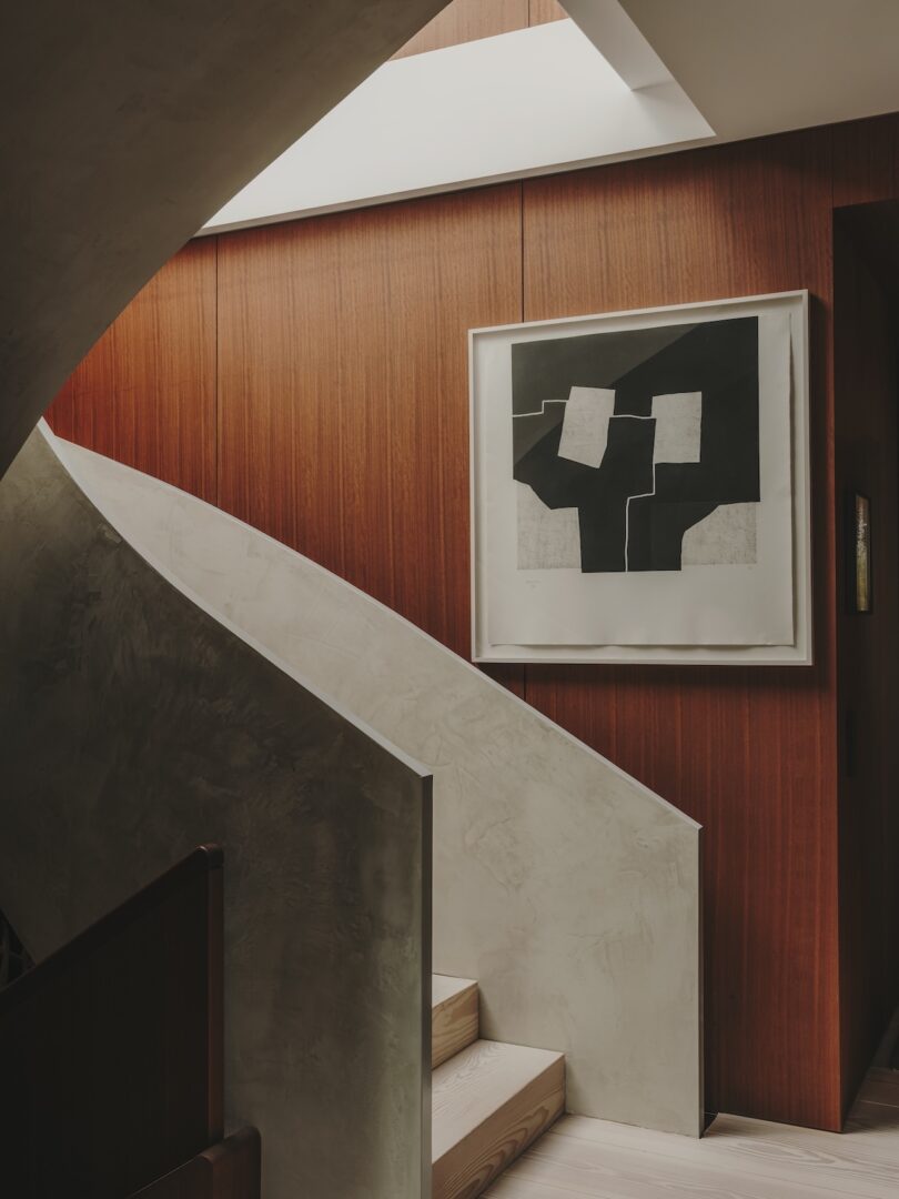 A curved staircase with a smooth, light-colored finish ascends beside a wooden wall featuring a framed abstract artwork in black, white, and gray tones.