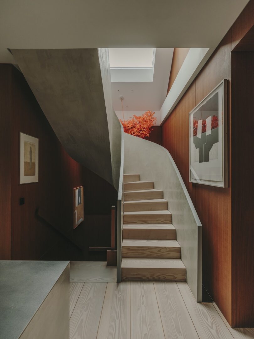 Minimalist staircase with light wood steps and a concrete banister, surrounded by wooden walls and modern artwork; skylight above and bright orange sculpture at the top.