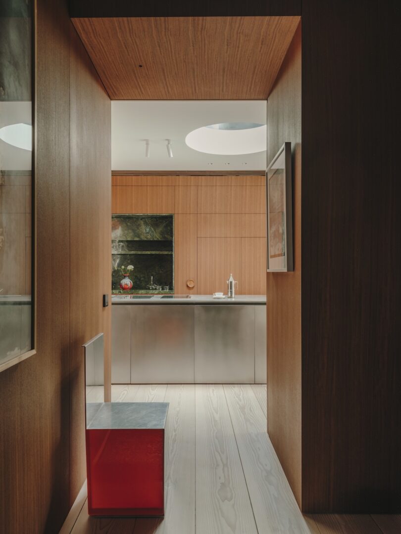 View of a modern kitchen with wooden walls, light wooden floor, stainless steel cabinets, and a red bench in the hallway. Natural light enters from a circular skylight above.