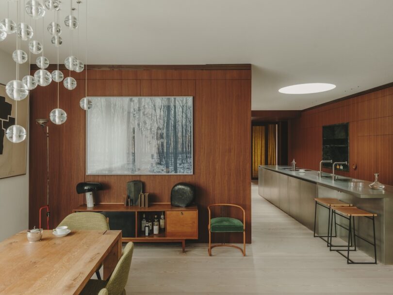 Modern dining and kitchen area with wood-paneled walls, minimalist furniture, a large forest art piece, and spherical hanging light fixtures.