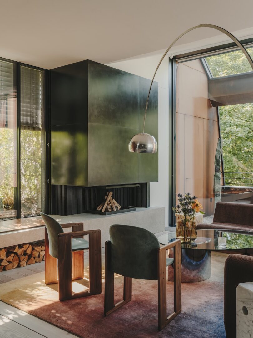 Modern living room with two green velvet chairs, a curved floor lamp, a wall-mounted fireplace, stacked firewood, and large windows overlooking greenery.