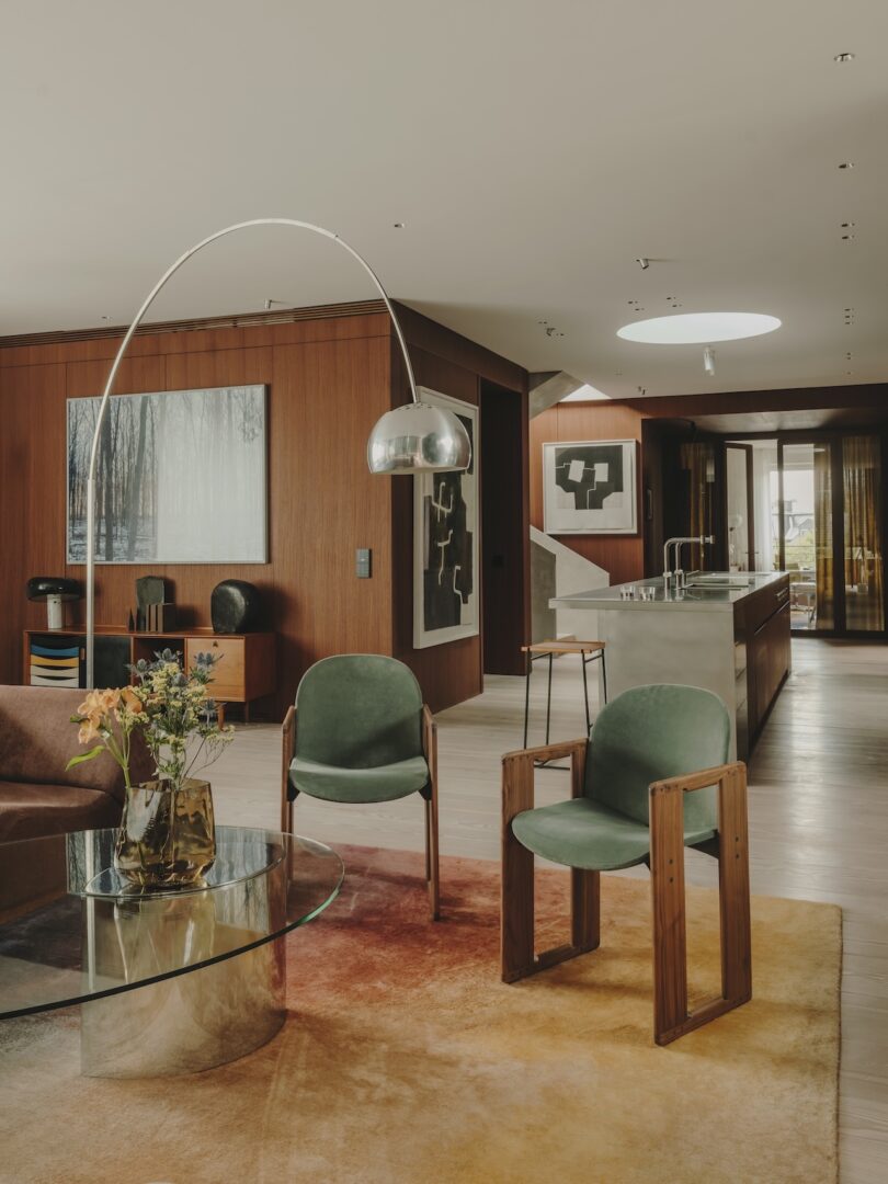 Modern living room with wood-paneled walls, green chairs, a glass coffee table, an arc floor lamp, and contemporary art, adjacent to an open kitchen with a minimalist design.
