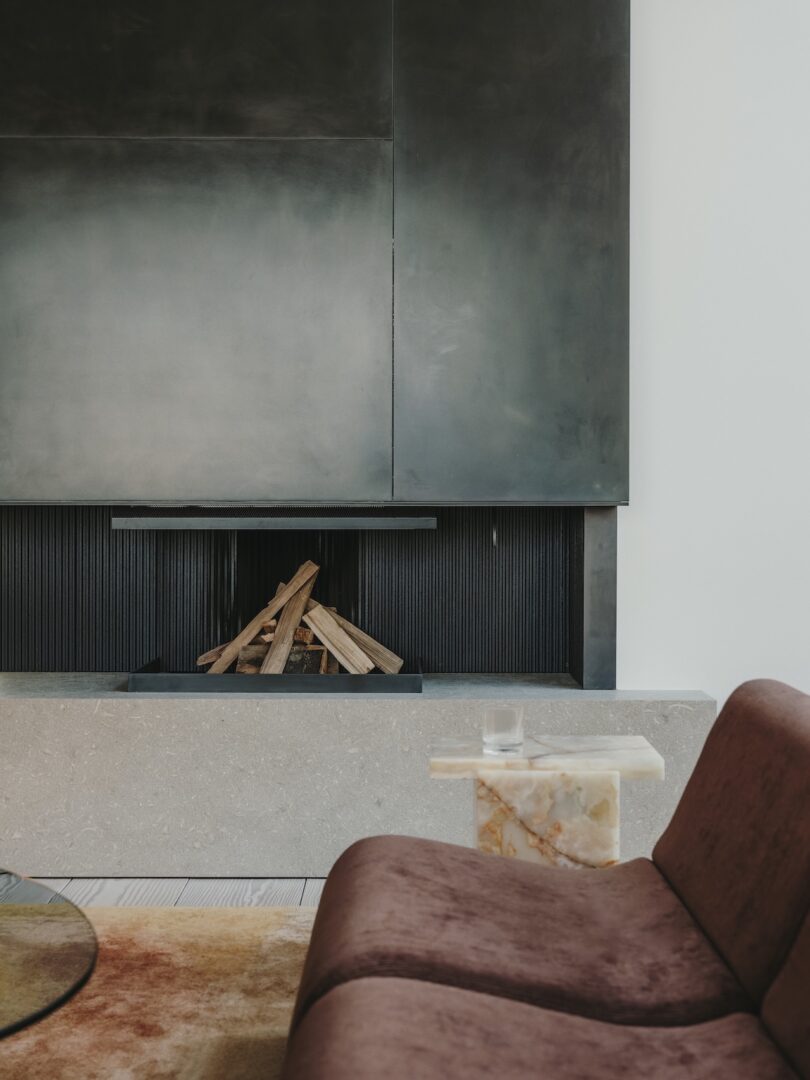 A modern living room with a dark metal fireplace, stacked firewood, a marble side table with a glass of water, and a brown velvet armchair.