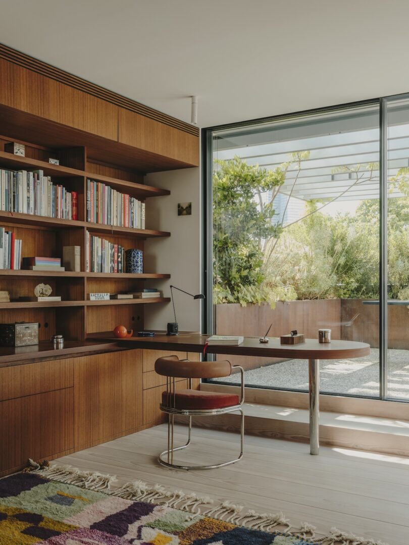 Modern home office with built-in wooden shelves, books, a desk with a chair, and large sliding glass doors opening to a garden patio with greenery outside.