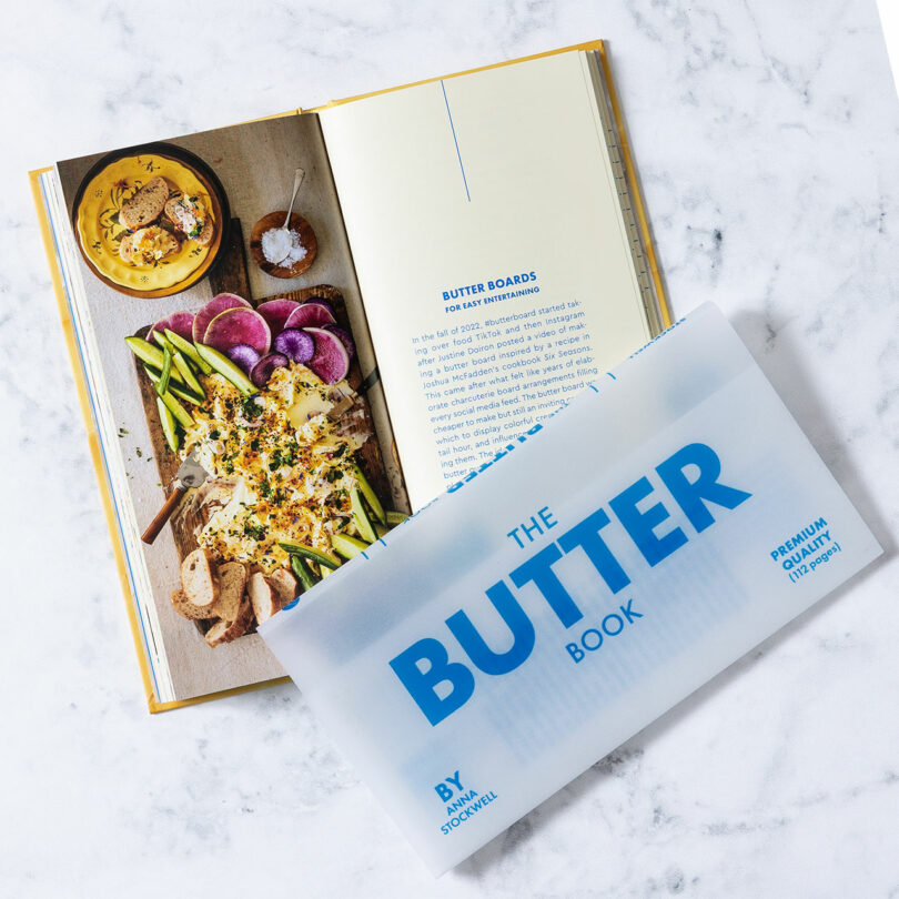 An open cookbook displays a recipe and a photo of a butter board with vegetables and bread, inviting you to experience the taste, with a translucent page labeled "The Butter Book" partially covering the book.